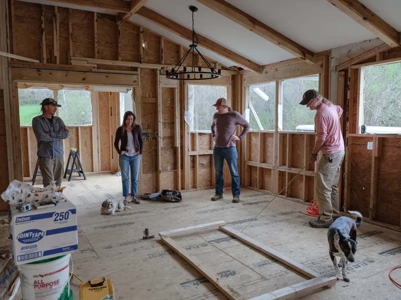 First image: A partially complete two-story house covered in Tyvek with stacks of lumber sitting on the ground. Second image: Horses feed on grass while trees begin to bloom. Third image: Four people smile inside of a wooden house in the process of being built.