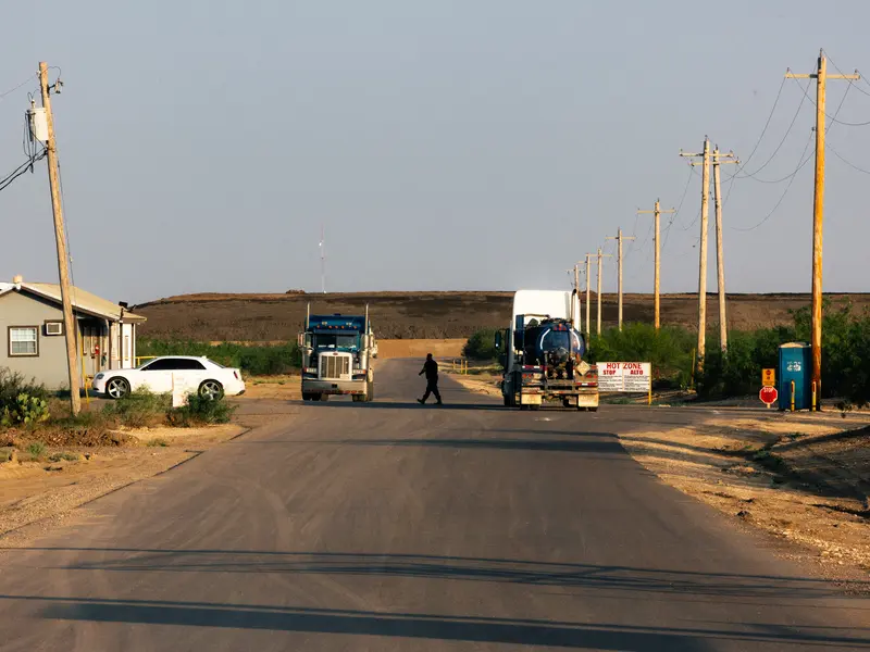 A man walks between two semitrucks on a road where only one small building is visible. A large, flat-topped hill made of darker soil stretches across most of the background and continues off the right side of the photo.