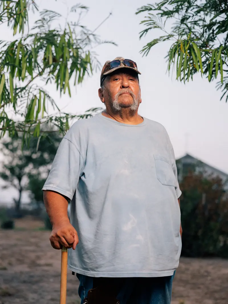 A man with a horseshoe mustache leans on a light wooden cane while standing outside. He wears a baggy blue T-shirt and a baseball cap with sunglasses resting on the brim.