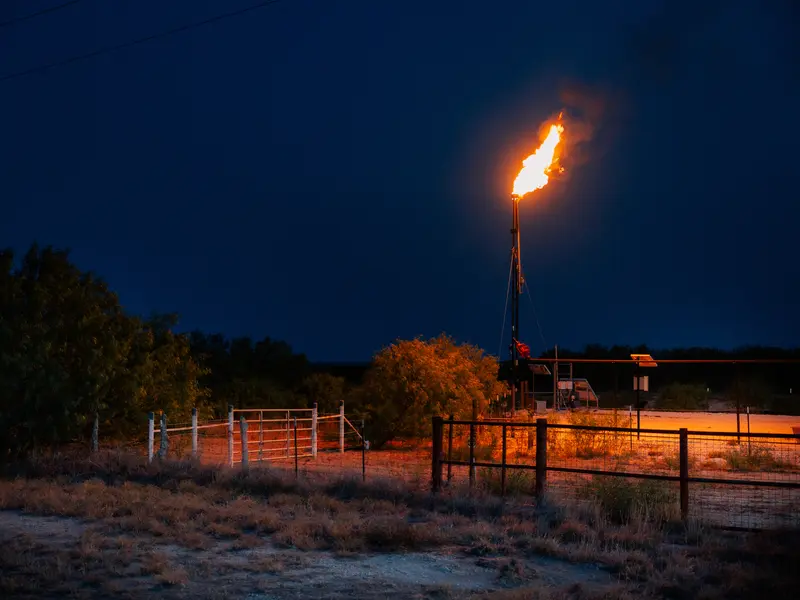 A jet of flame rising from a tall pipe casts an orange glow over an area with fences and brush.