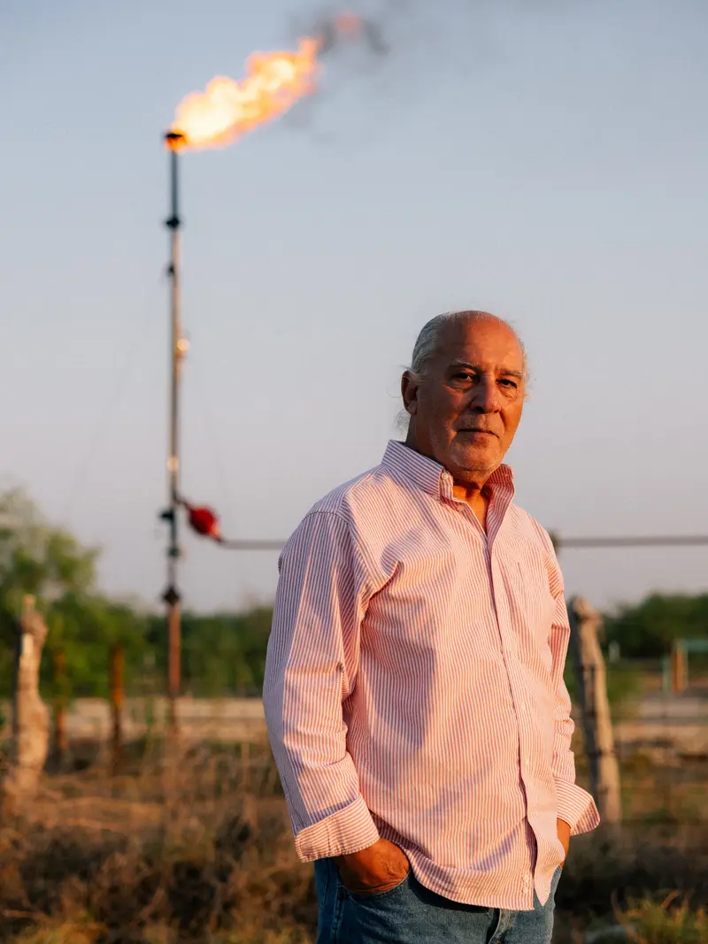 A man in a button-down shirt with thin pink and white vertical stripes stands with his hands in his jeans pockets. Behind him, flame jets from the top of a pipe that stands straight up in the air.