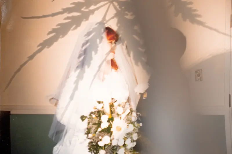 An old photograph showing Sutton-Schulman with a veil over her red hair, wearing a white wedding dress and holding a bouquet. She is looking at her husband, whose face is obscured by a shadow falling on the photograph.