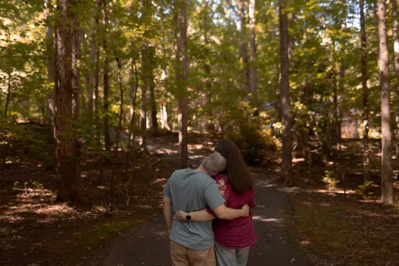 Two people standing in the woods with their backs to the camera. L has short gray hair and is wearing a blue T-shirt. Sutton-Schulman has dark hair flowing to the bottom of her shoulderblades and is wearing a red shirt with flowers on the sleeve. L is resting his head on Sutton-Schulman’s shoulder and the two have their arms around one another.