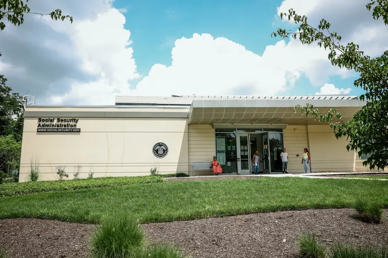 A beige building with big letters on the front reading “Social Security Administration” with a few people outside the front door.
