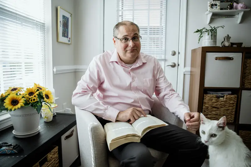 A man sits on a chair in a brightly lit room with a book open on his lap. A white cat is in the lower right-hand corner.