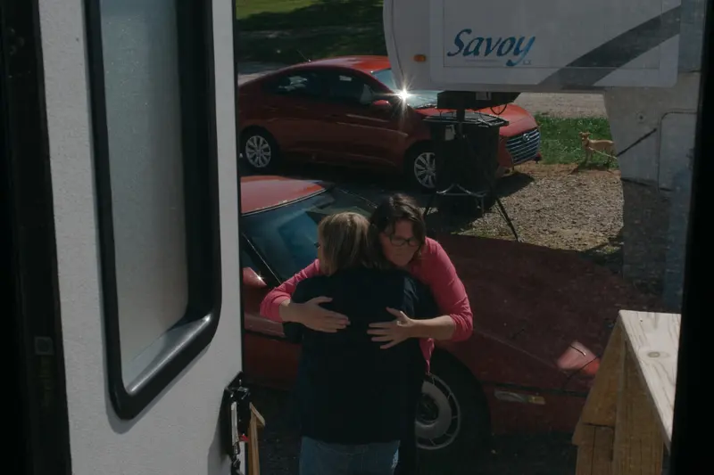 Two women embrace at the bottom of a wooden staircase in front of two red cars.