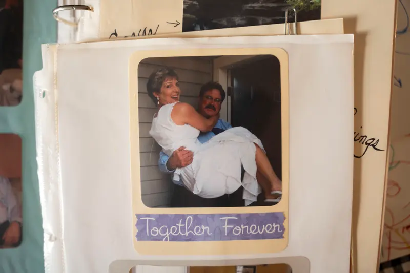 A scrapbook photo shows a man with a pushbroom mustache picking up a woman in a white dress with the caption “Together Forever” underneath. Both are smiling. The woman has short blonde hair and wears white flip-flops with her dress. The man wears a long-sleeve blue shirt, dark pants and glasses.