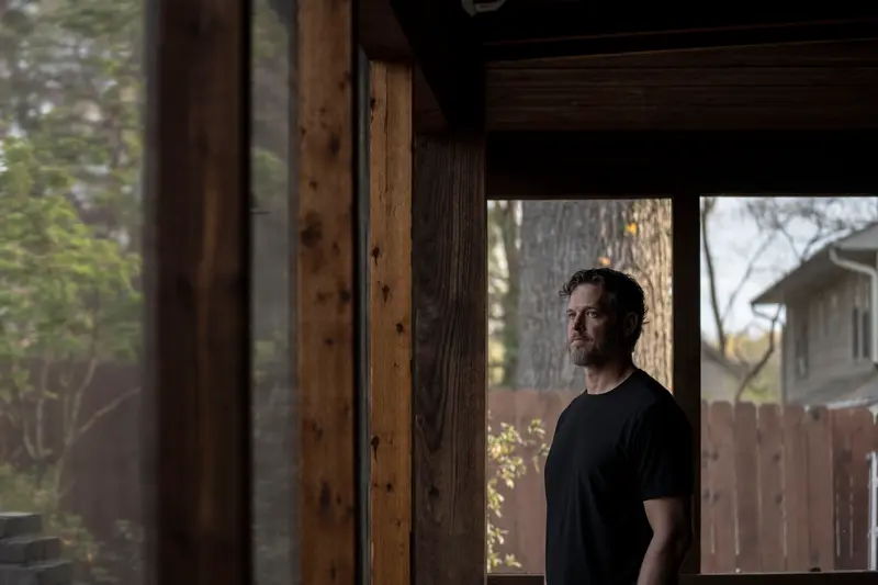 A man wearing a black shirt stands inside a screened-in porch looking out toward a woodsy backyard