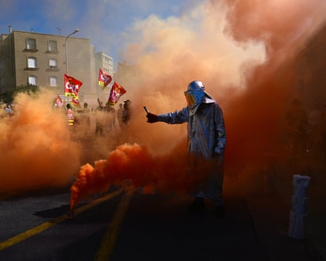 A man in a heat-protective suit holding an orange flare as others burn on the ground