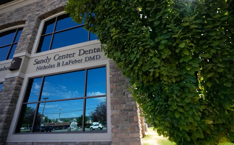 A large green bush partially covers a building with a sign that reads: Sandy Center Dental, Nicholas B. LaFeber DMD. The windows reflect a blue sky and shopping center parking lot.