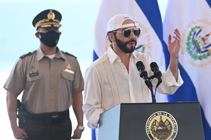 Bukele speaks at a podium wearing a backward white baseball hat and aviator glasses.