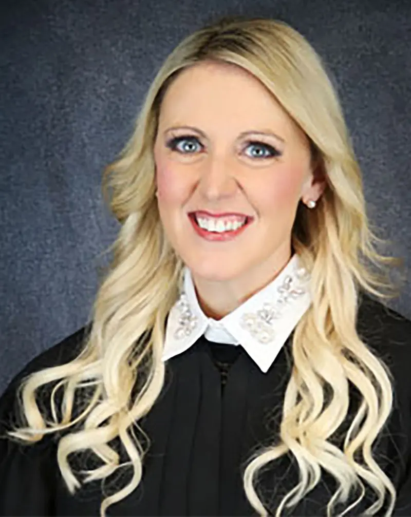 A woman with long, wavy blond hair and blue eyes smiles at the camera. She is wearing a white shirt with a collar and a black judicial robe.