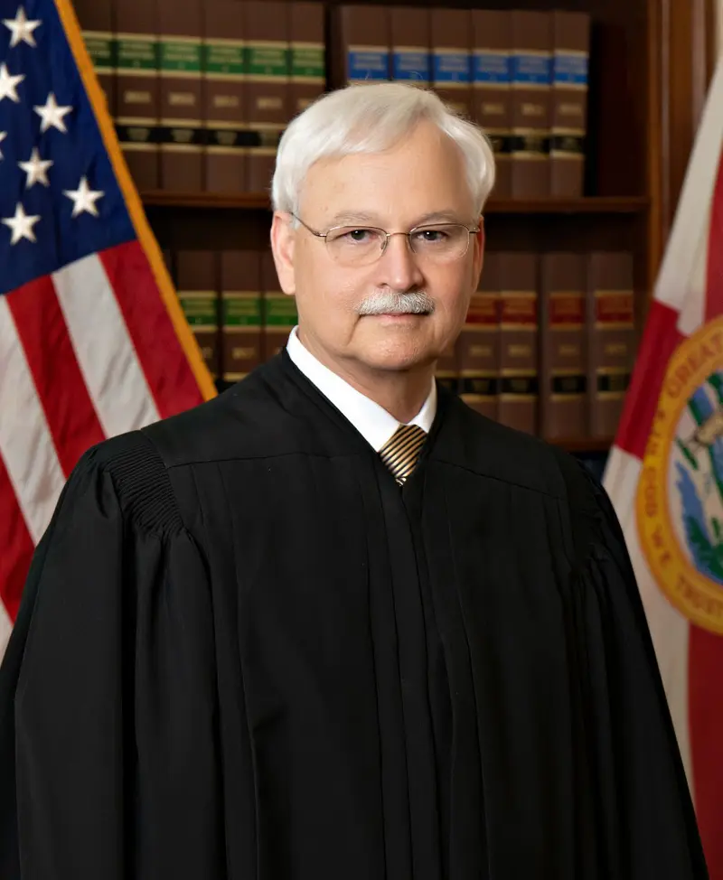 A man with glasses, a white mustache and white hair wears a tie and a black judicial robe and is standing in front of the flags of the United States and Florida and a bookcase full of legal books.