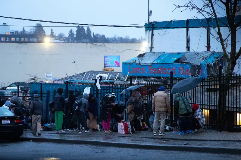 A line of people with shopping bags, umbrellas and coats sit and stand along a black fence topped with razor wire, next to a building with the sign “Lam’s Seafood Market.”