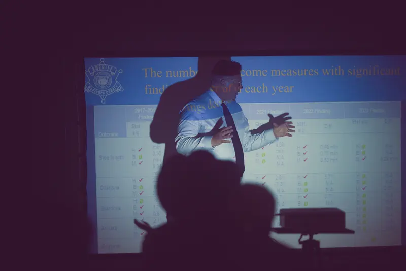 A man in a white shirt and tie gestures with his hands apart in front of a projector with a data table on the screen behind him.