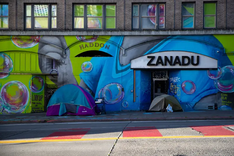 Two tents sit on a sidewalk in front of a wall with a mural in bright blue and green.