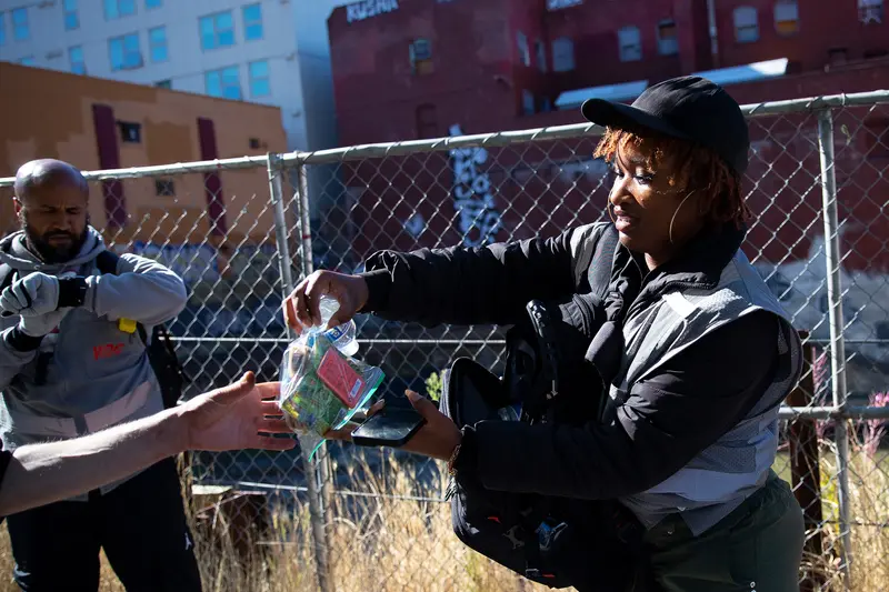 A woman wearing hoop earrings and a baseball cap hands a ziplock bag and a small water bottle to someone out of frame. A man checks his watch behind them. They are standing in front of a chain-link fence, overgrown grass and buildings tagged with graffiti.