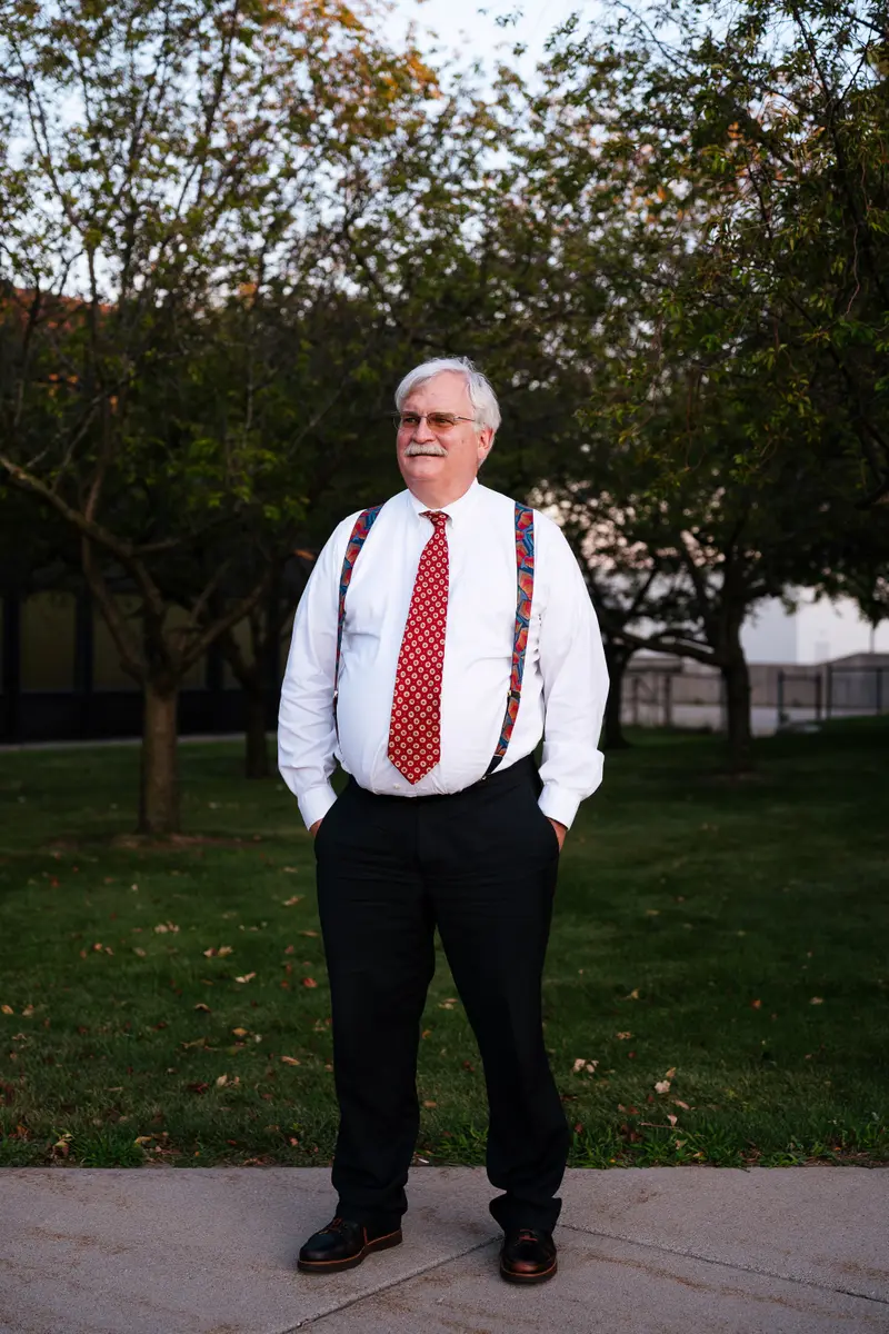 Dr. Mert Aksu, dean of the University of Detroit Mercy’s dental school, poses for a portrait near a park with grass and trees while wearing a white dress shirt, red tie, suspenders and black slacks.