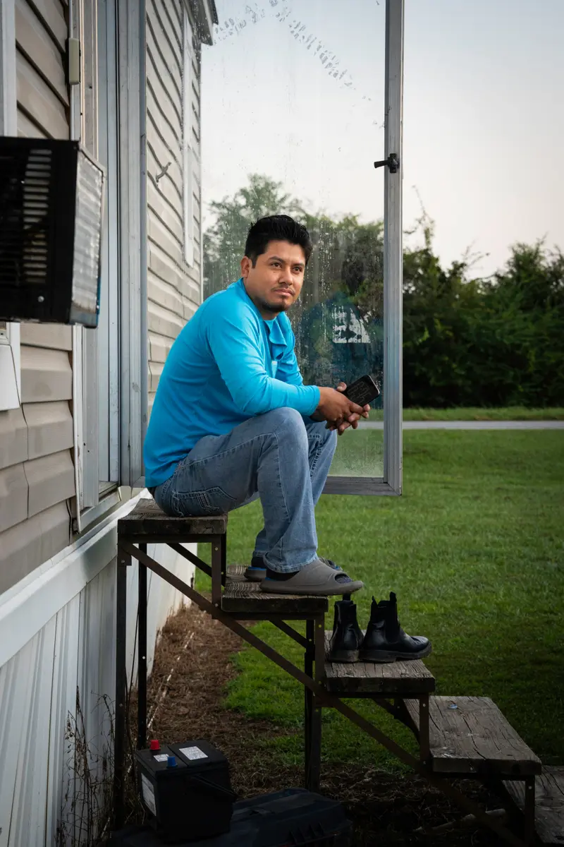 A man with a small goatee seated on the steps of a single-wide home, wearing a blue shirt, jeans and a pair of sandals.