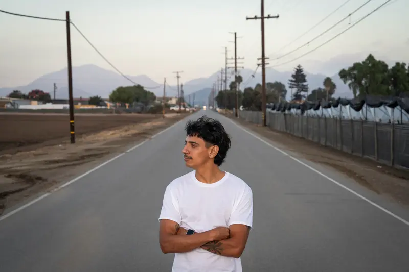A man with a mustache wears a white T-shirt and stands with his arms crossed on an empty road.