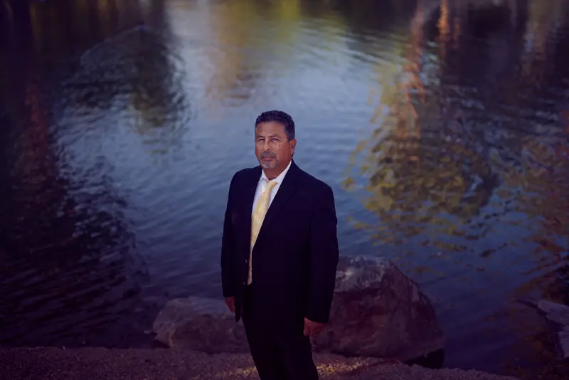 Raul Piña, a man with dark hair and a salt-and-pepper goatee, stands in front of a small body of water wearing a suit and a yellow tie.