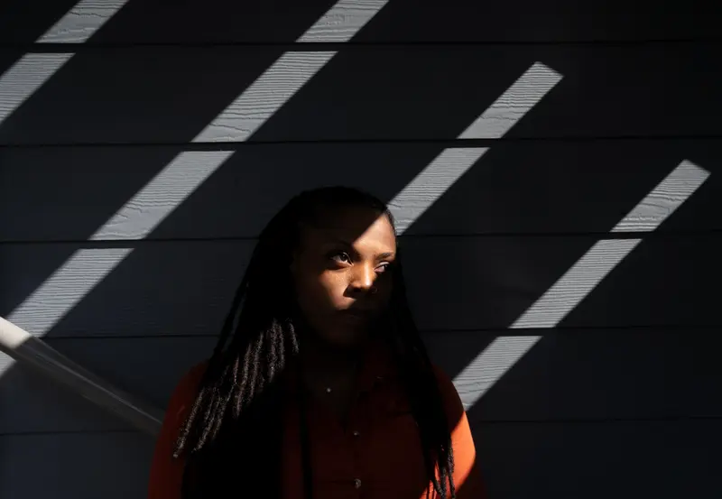 A woman stands in the dark with diagonal slats of light falling on her face and arm.