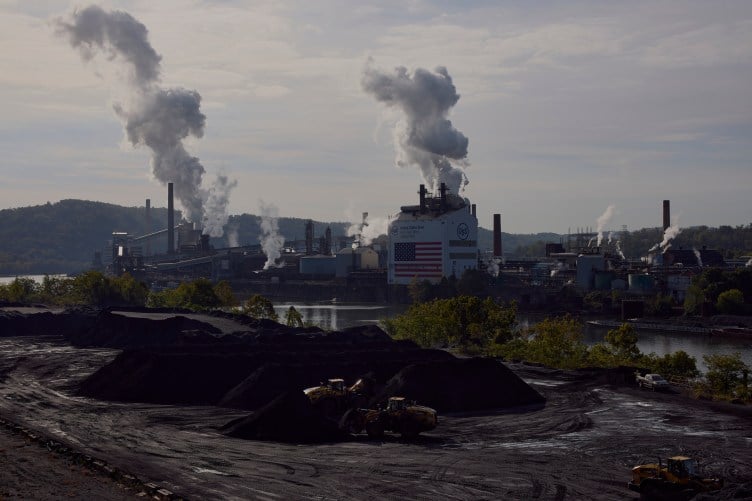 Yellow trucks navigate large piles of black sandy coal in front of a river. Across the river is a large industrial complex with thick steam billowing from several buildings, one of which has an American flag painted on the side. 