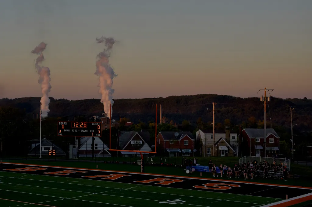 Two large columns of steam rise up in the background behind a high school football field at sunset.