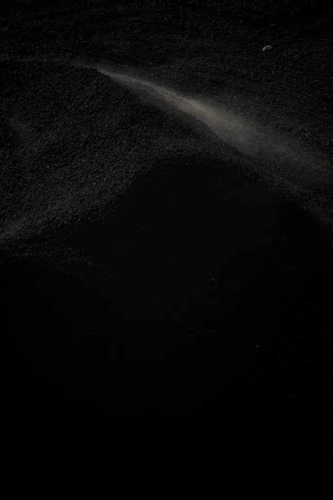 A single beam of light illuminates the top edge of a large pile of black sandy coal.