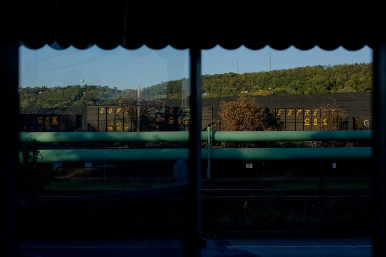 Train cars and large green pipes running along a road, framed by a residential window with scalloped window treatments at the top.