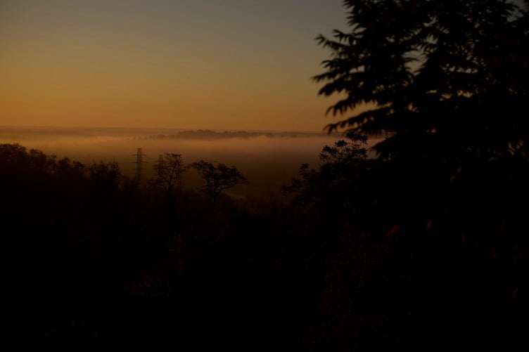 A wide aerial landscape view of a dark valley filled with smog, surrounded by trees, glowing in warm early-morning light.