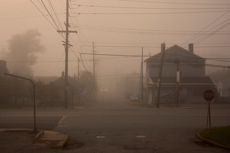 Heavy smog clouds the air in a deserted-looking working class residential intersection with many power lines running overhead. A pair of headlights glow weakly through the haze.