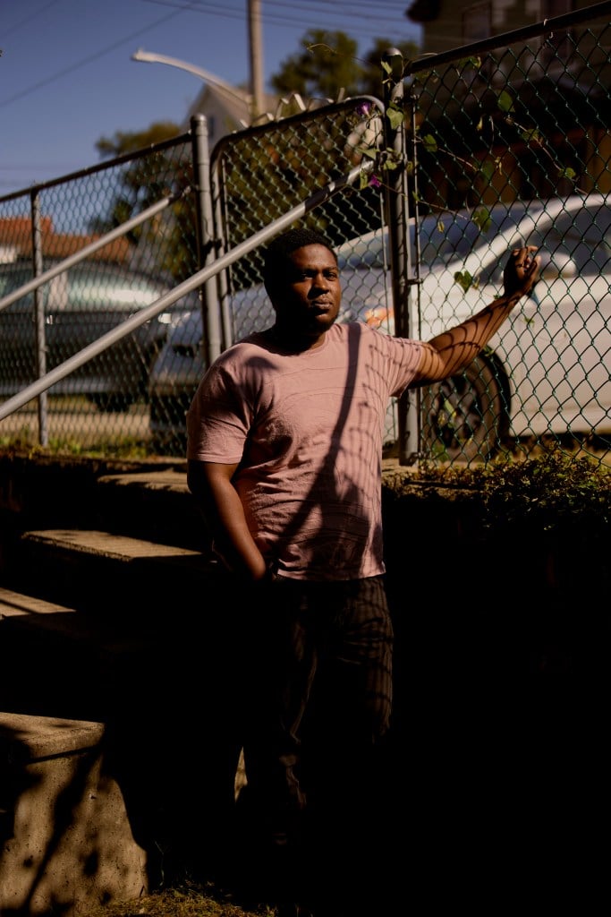 Ansari, standing on a residential stoop, leans against a chain-link fence while looking at the camera, his face illuminated by bright dramatic sunlight.