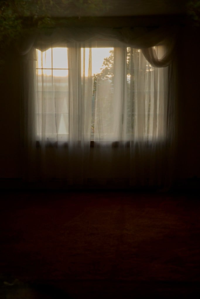 A yellow glow suffuses the window of a dark residential living room with sheer curtains.