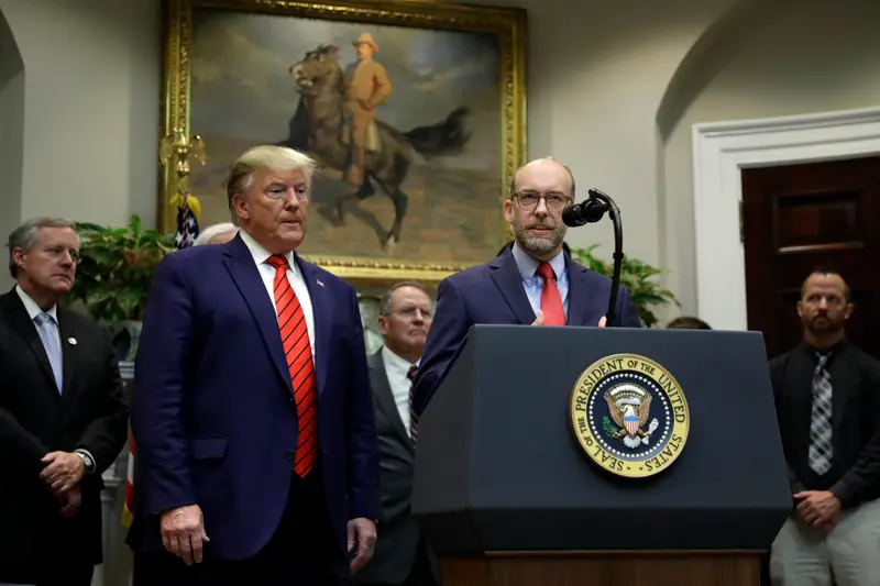 Russ Vought stands at a podium with Trump beside him.