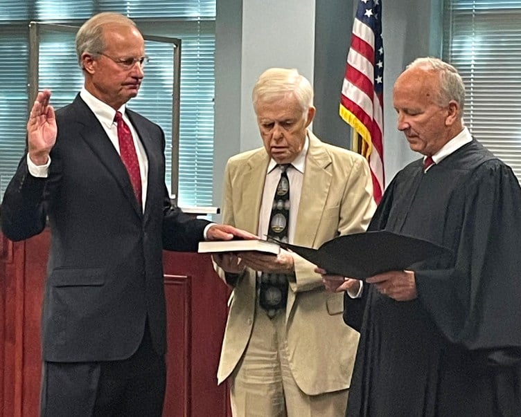 Van der Vaart, a man with glasses and gray hair wearing a suit and tie, raises his right hand while resting his left on a book held by another man. Newby, in judges’ robes, reads from an open folder.