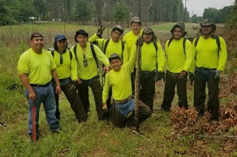 A smiling group of men wearing neon yellow shirts, hats and long pants pose for the camera in a grassy area. One man kneels and holds up_ _one end of a large snake with his arm straight in the air while the other end rests on the ground.