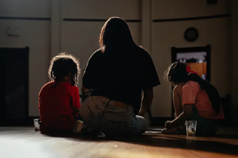 A woman, seen from behind, plays with two children on the floor of a dark room.