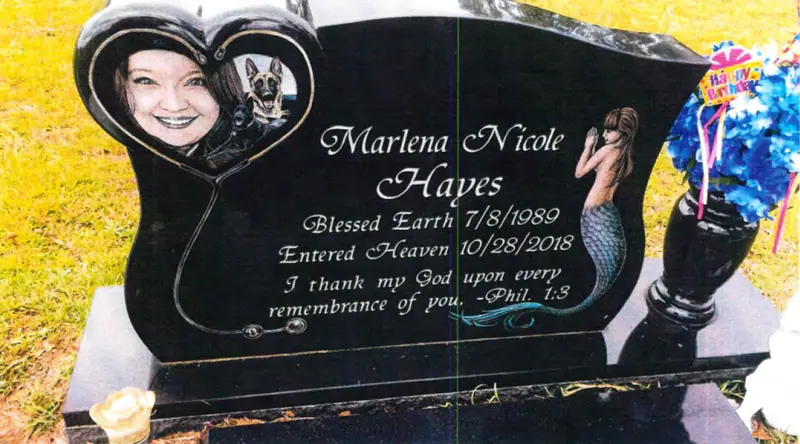 A black headstone with the words “Marlena Nicole Hayes. Blessed Earth 7/8/1989. Entered Heaven 10/28/2018. I thank my God upon every remembrance of you. —Phil. 1:3.” A mermaid and a heart with a smiling woman and a German shepherd also appear on the headstone. A vase of blue flowers with the words “Happy Birthday” sits on the grave.