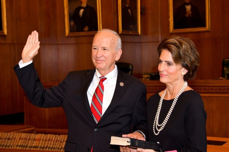 A man in a suit and tie with white hair rests his hand on a Bible held by a woman with brown hair wearing a black dress and a string of pearls. They are standing in a wood-paneled room lined with framed portraits and law books.