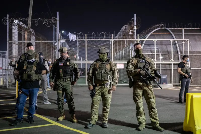 Masked agents stand outside of a detention center surrounded by metal fences and barbed wire.