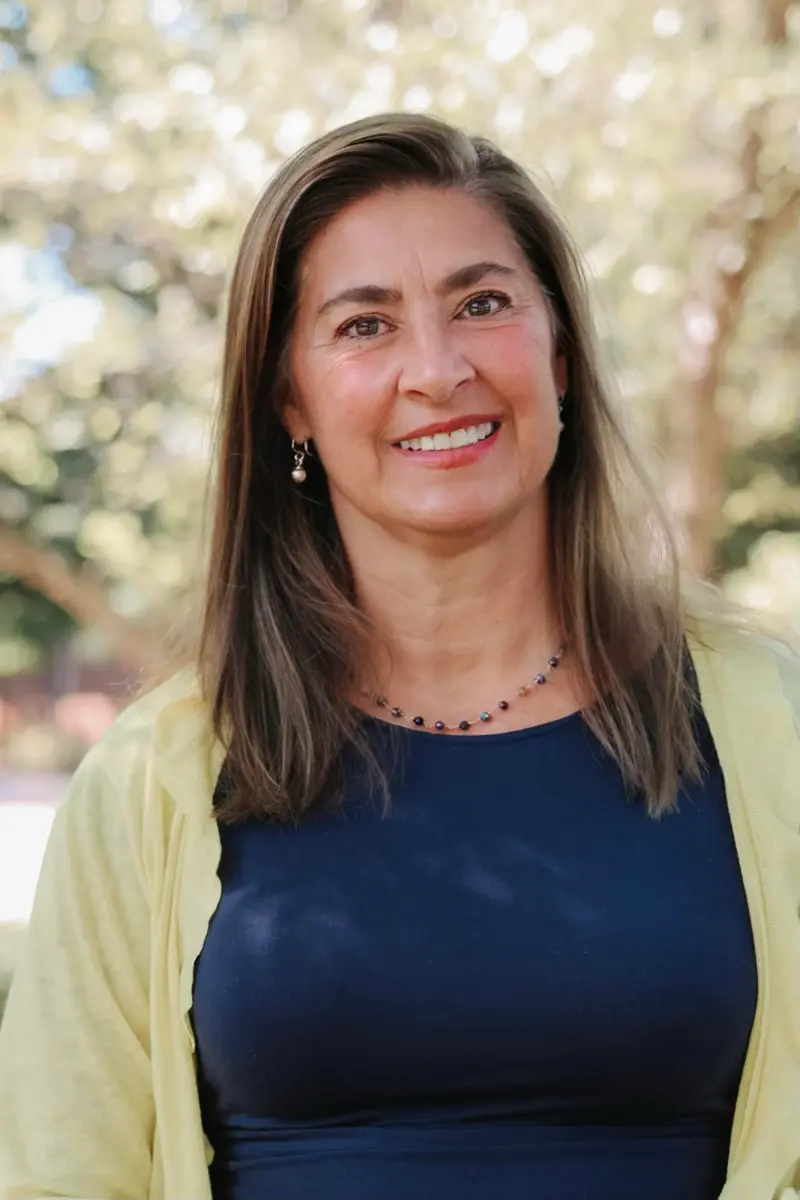 A woman with shoulder length brown hair, a yellow cardigan and blue shirt smiles at the camera.