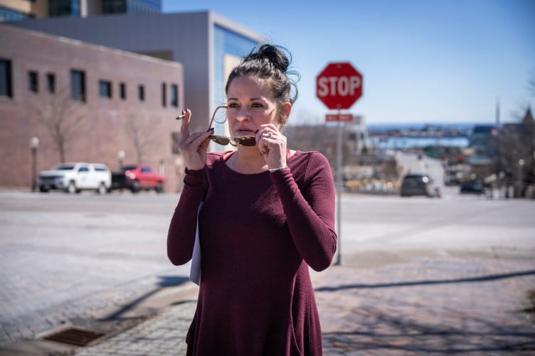 A woman with her hair in a bun, wearing a maroon outfit, adjusts her glasses while holding a cigarette in one hand as she stands on a street corner in a downtown urban environment on a sunny day.