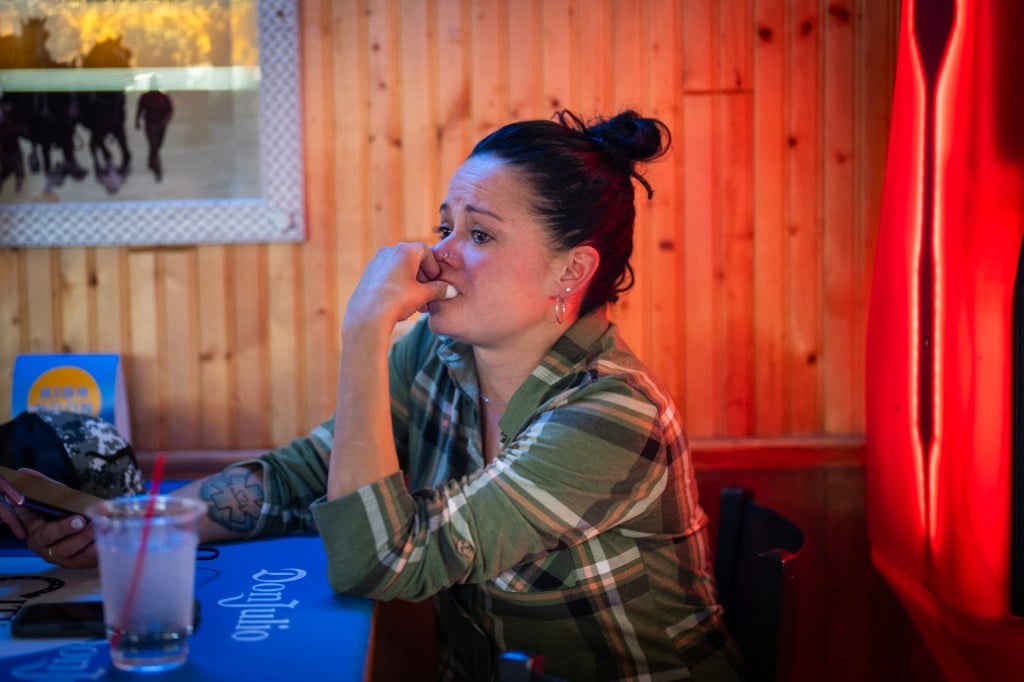 A woman with black hair in a bun, wearing a green shirt, cries at a blue table in a dimly lit bar.