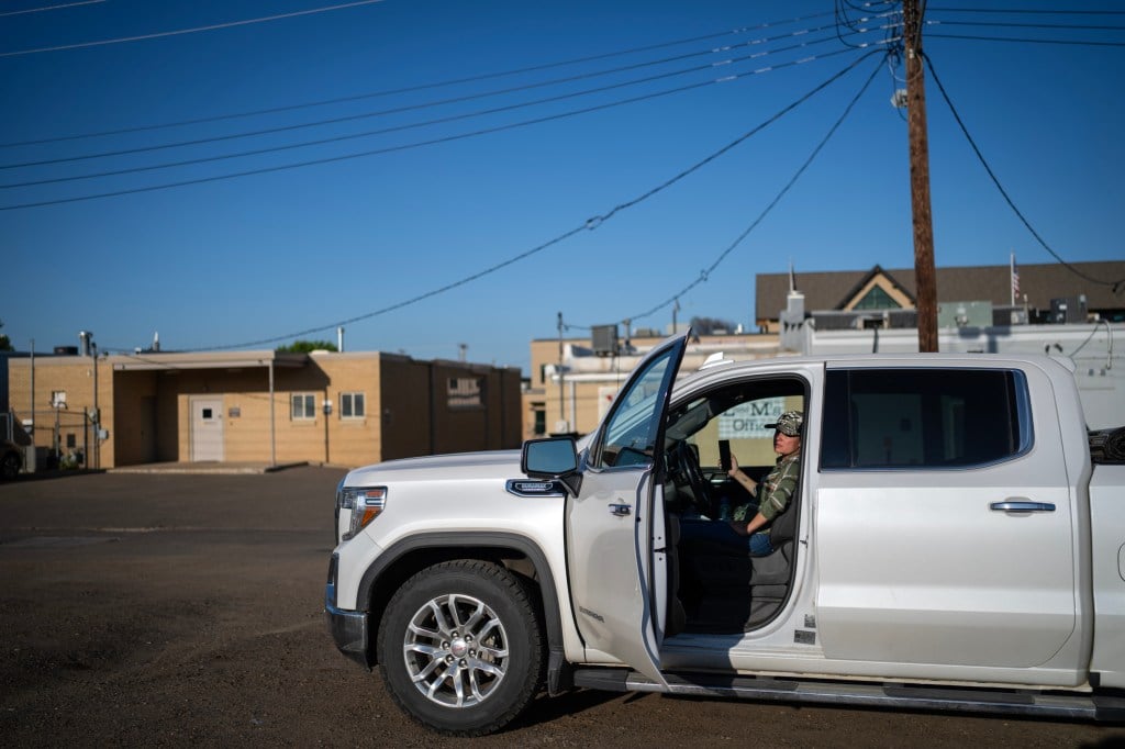 A woman wearing a green shirt and holding a cellphone sits in the cab of a white pickup truck in a parking lot behind a section of yellow buildings.