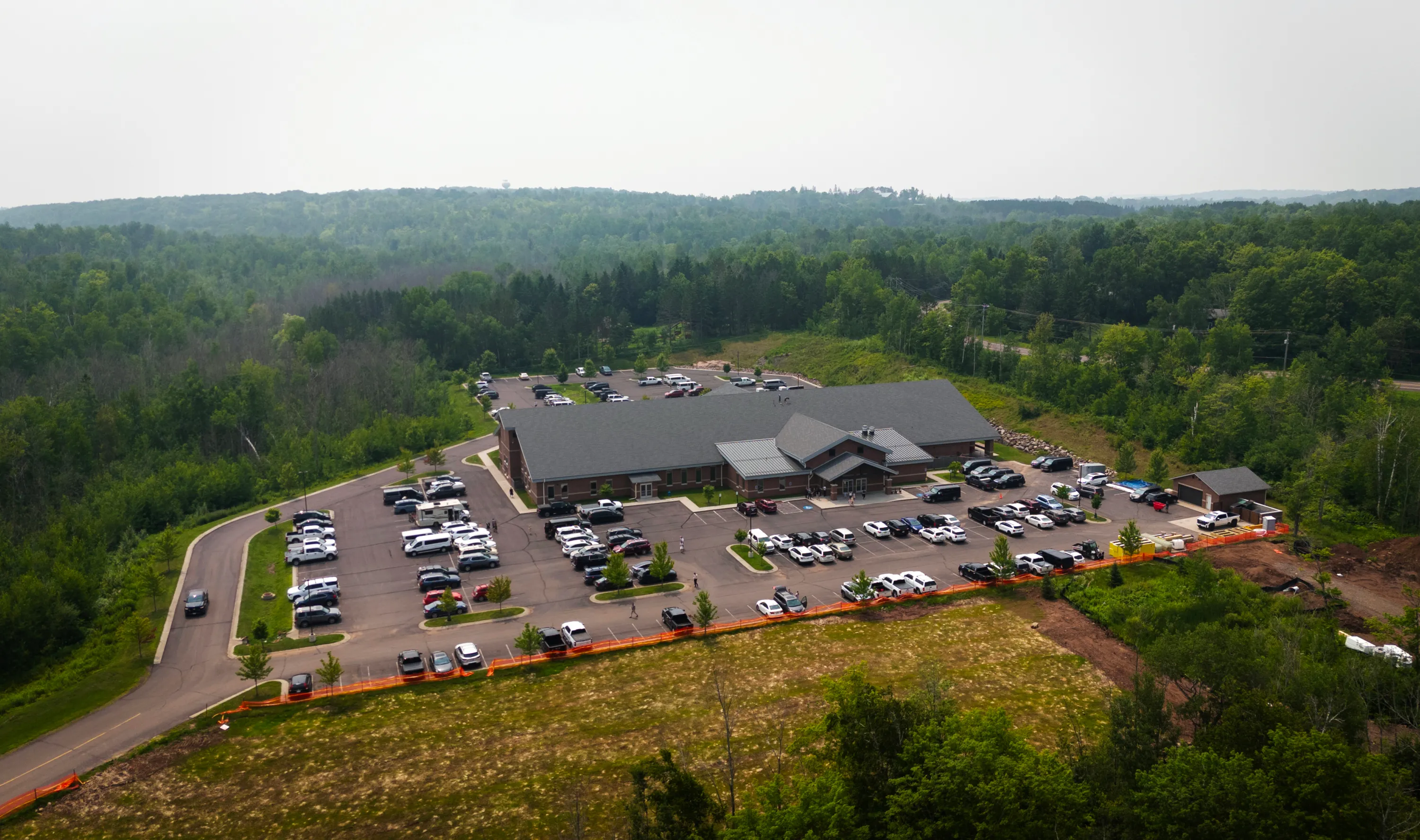 A large church building surrounded by a parking lot filled with cars and dense woods on a foggy morning.