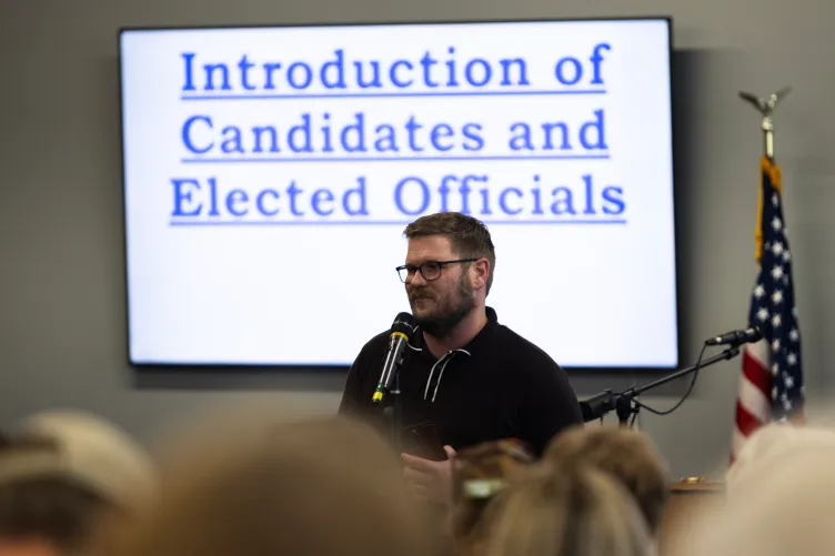 A bearded man in a black polo speaks in front of a screen that says “introduction of candidates and elected officials.”