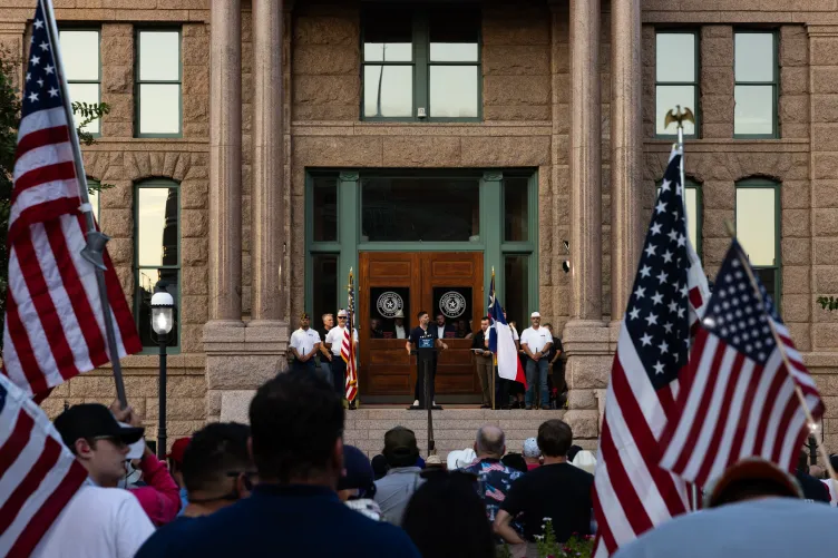A man, seen from the back of a crowd, stands on the steps of a stone building behind a small lectern. Multiple people in the crowd are waving large American flags.