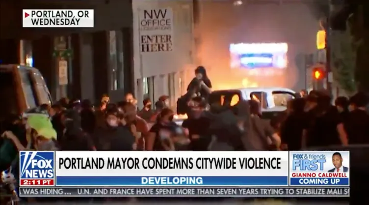 A crowd of people on a street in front of smoke and fire with a chyron that says: “Portland mayor condemns citywide violence.”