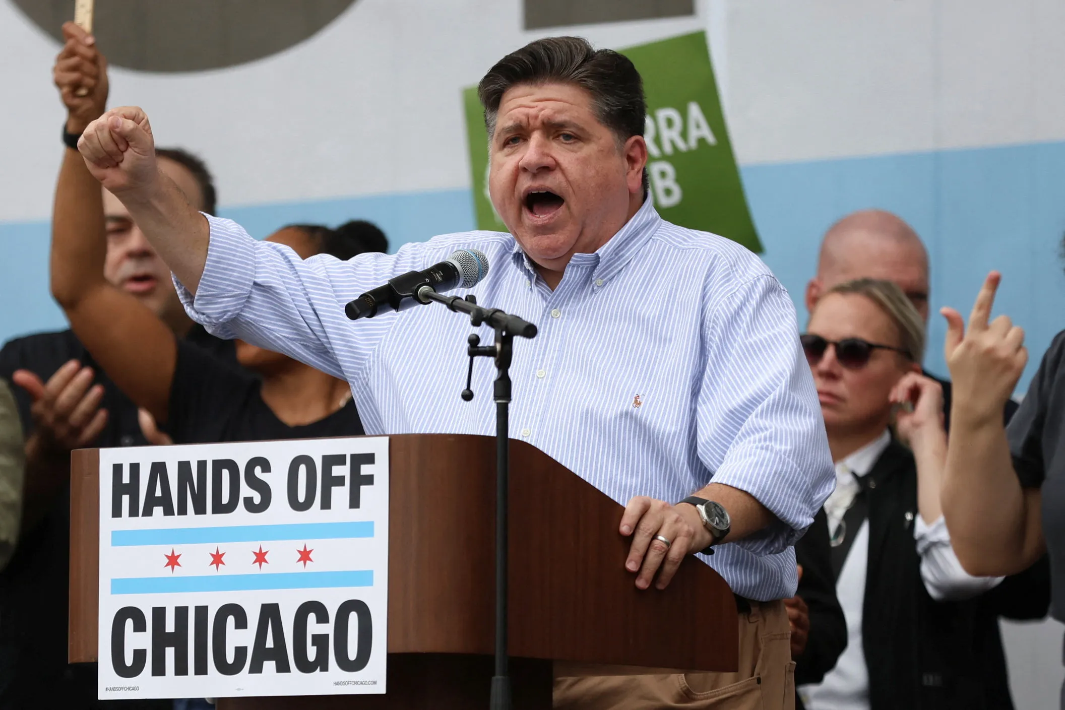 A man stands behind a lectern raising his fist. A sign on the lectern reads, “Hands off Chicago.”